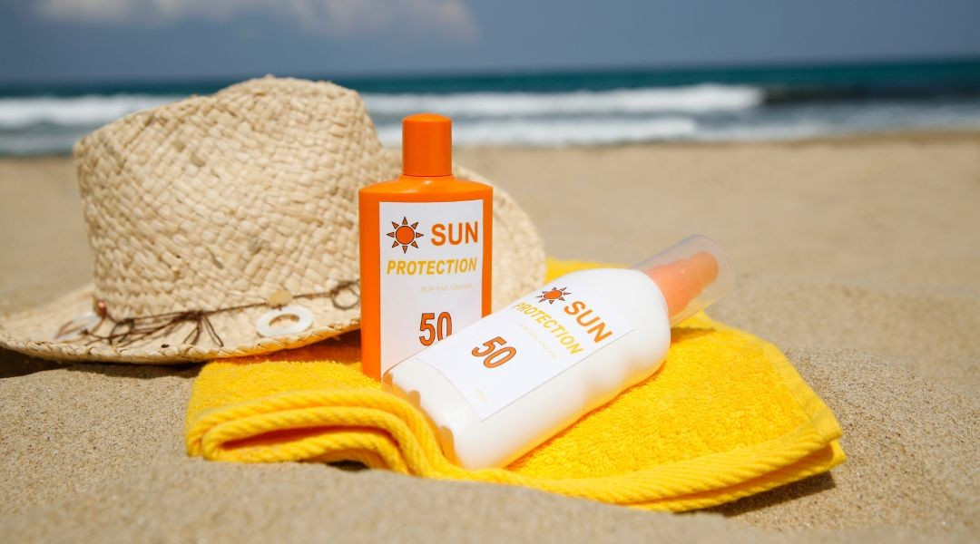 Two bottles of sun tanning lotion with a towel and sun hat on the beach.